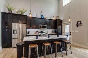 A modern kitchen with dark cabinets, stainless steel appliances, a central island with stools, hanging pendant lights, and decorative items on the counter.