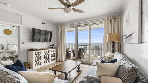 A spacious living room with a large TV, a ceiling fan, and a view of the ocean through a sliding glass door. - Emerald Isle 506 - Beautiful beachfront condo! Front row chairs included! (Panama City Beach)