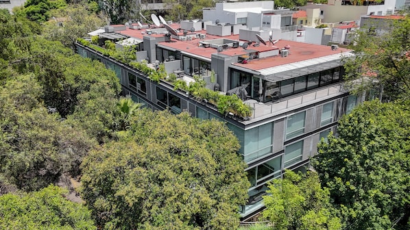 Un edificio moderno de varios pisos con una terraza en la azotea, rodeado de abundante vegetación.