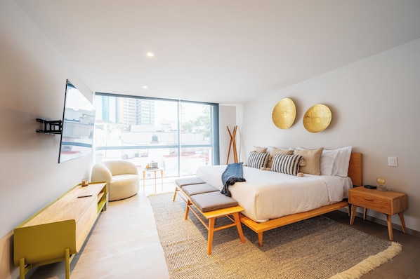 A modern bedroom with a large bed, a desk, a chair, and a view of the city through large windows. - Capitalia Parque Lincoln (Mexico City)
