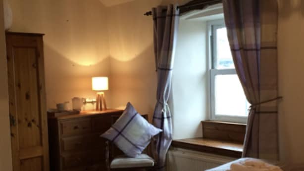 A bedroom with a wooden wardrobe, a desk with a lamp, a chair, a window with curtains, and a bed with a folded towel on it. - Herdwick Inn (Penrith)