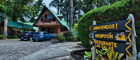 A sign with 'RECEPTION' and 'RESTAURANTE LAS GALAS' directions, a building with a green roof, and parked cars.