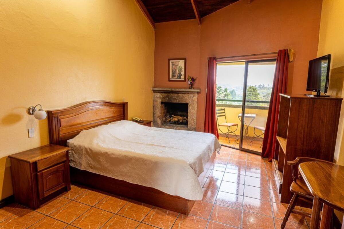 Un dormitorio con cama, mesita de noche, silla de madera, televisor, chimenea y balcón con mesa y sillas.