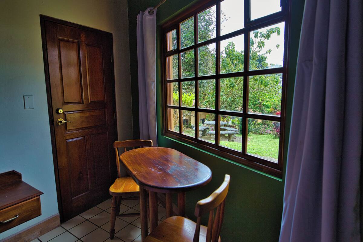 Una habitación con una mesa de madera y dos sillas, una ventana que da a zonas verdes y una puerta cerrada.