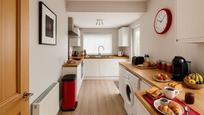 Eine moderne Küche mit einer zentralen Kochinsel, einer Waschmaschine, einem roten Mülleimer und einer Frühstücksecke mit Brot, Früchten und Kaffee.