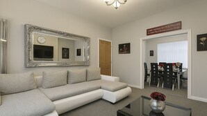 Ein Wohnzimmer mit einer Eckcouch, einem Couchtisch mit einer Vase, einem großen Spiegel und einem Essbereich mit Tisch und Stühlen.