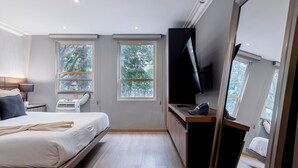 Un dormitorio moderno con cama, escritorio y televisión. Dos ventanas permiten la entrada de luz natural.