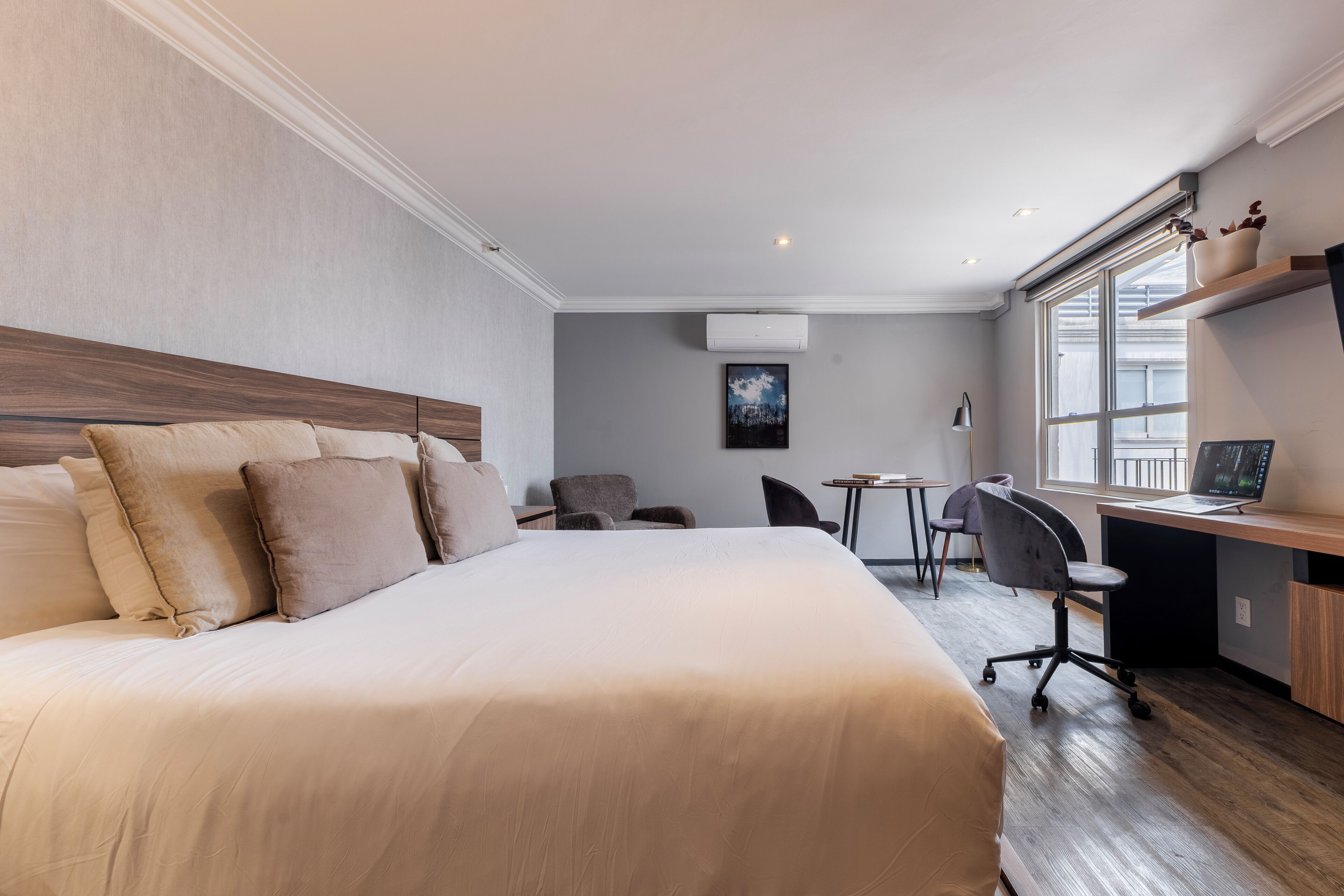 A modern hotel room with a large bed, a desk, and a chair. There is a window providing natural light and a view of the outside.
