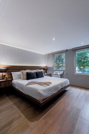 A modern bedroom with a large bed, wooden headboard, bedside tables with lamps, and large windows with a view of trees. - Capitalia Obelisk Polanco (Mexico City)