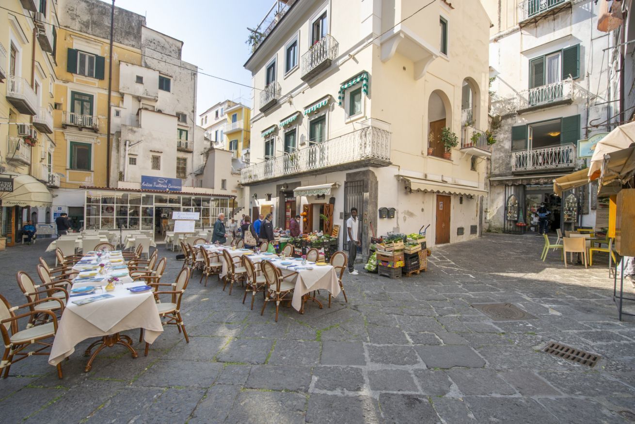 An outdoor dining area with tables and chairs, surrounded by buildings with balconies and shops.