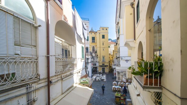 Uma rua estreita com edifícios em ambos os lados, mesas ao ar livre e algumas pessoas caminhando.