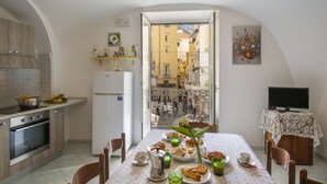 Uma sala de jantar com mesa posta para o café da manhã, uma cozinha com eletrodomésticos modernos e vista para a rua através de uma porta aberta.