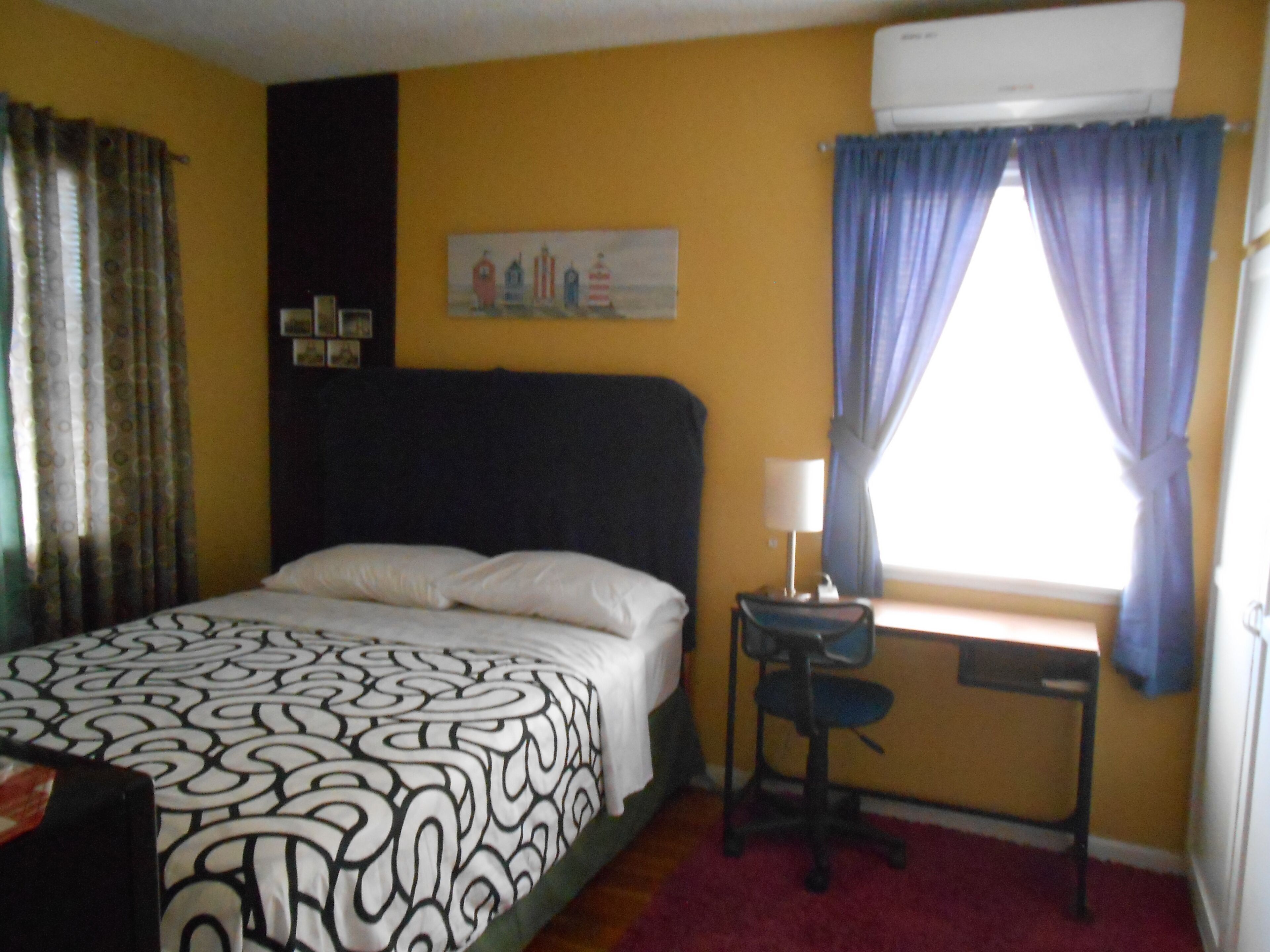 A hotel room with a bed, a desk with a chair, a window with curtains, and a painting on the wall.