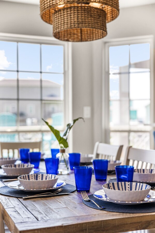 Une table à manger dressée avec de la verrerie bleue et des bols à rayures, un luminaire en fibres tressées et une vue dégagée sur l’extérieur grâce à de grandes fenêtres.