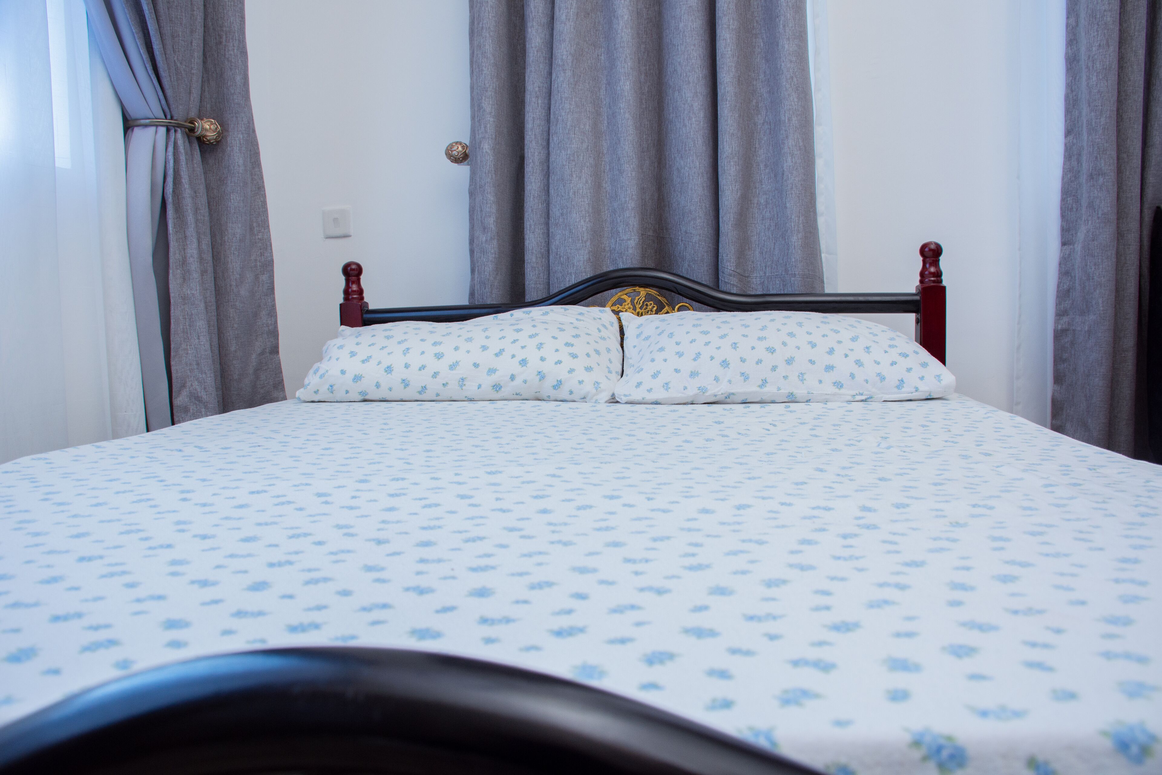 A neatly made bed with a floral-patterned bedspread and matching pillows, a wooden headboard, and grey curtains.