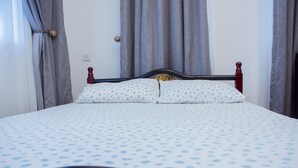 Un lit bien fait, avec une couvre-lit à motifs floraux et des coussins assortis, une tête de lit en bois et des rideaux gris.