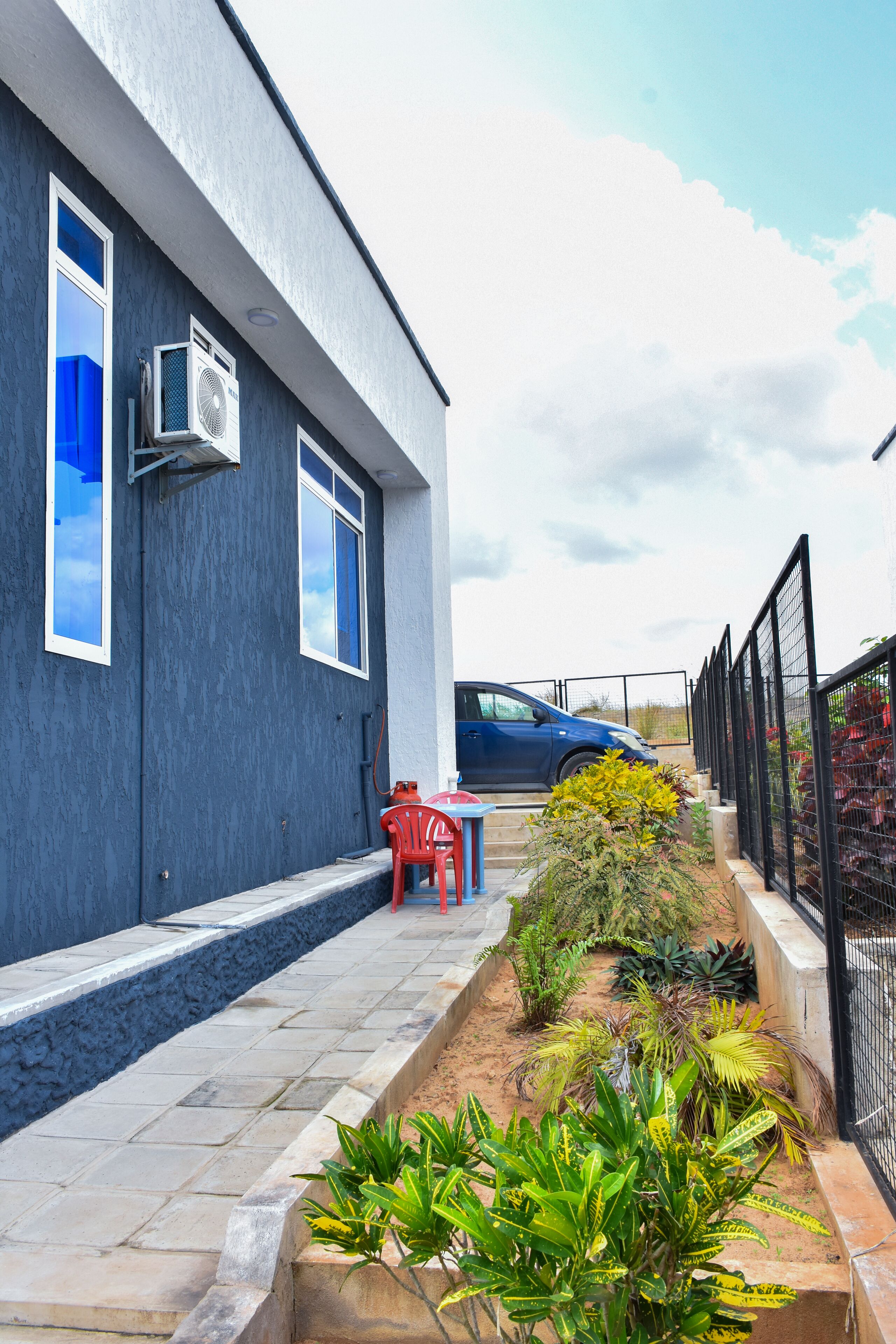 Un edificio moderno con fachada azul, un camino empedrado y un jardín con diversas plantas.