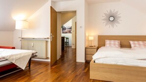 Habitación de hotel con dos camas, suelo de madera, radiador, lámpara de pared y puerta que da a un comedor.
