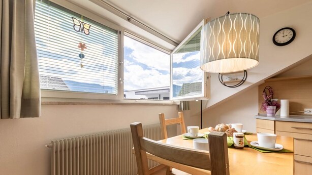 Un coin repas comprenant une table, des chaises, une fenêtre avec des stores, un radiateur et une lampe suspendue.