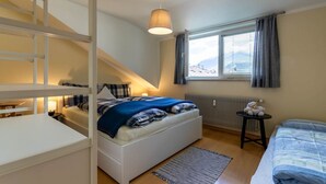 Une chambre avec un lit superposé, un lit simple, une petite table sur laquelle est posé un ours en peluche, une fenêtre avec des rideaux et un luminaire suspendu.