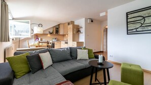 Un salon moderne avec un canapé d’angle gris, une petite table ronde et un fauteuil vert. On aperçoit une cuisine en arrière-plan, avec des meubles en bois et un coin repas.