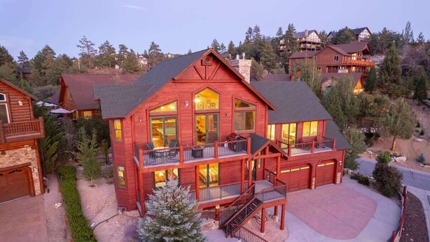 A large, multi-story wooden house with a prominent balcony, a driveway, and a well-maintained lawn. - Windsong Retreat | 3X2 PROMO | Lake Views. Hot tub (Big Bear Lake)