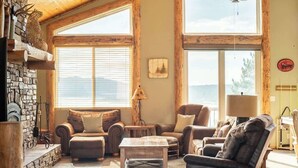 A cozy living room with a stone fireplace, leather sofas, a coffee table, and large windows offering a view of the outdoors. - Windsong Retreat | 3X2 PROMO | Lake Views. Hot tub (Big Bear Lake)