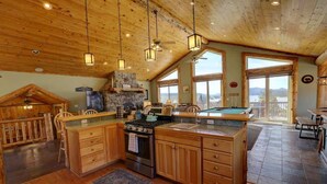 A spacious kitchen with wooden cabinets, a central island, and a stone fireplace. - Windsong Retreat | 3X2 PROMO | Lake Views. Hot tub (Big Bear Lake)