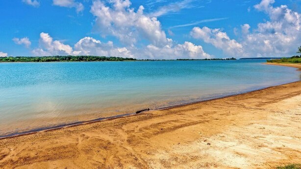 A beach with clear water, a sandy shore, and a distant tree line under a blue sky with clouds. - Best kitchen in Texoma! New! Playroom! (Mead)