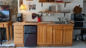 Una cocina compacta con gabinetes de madera, un refrigerador empotrado, un lavamanos y un microondas. Se aprecia una pared de piedra y un techo con vigas de madera.