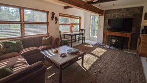 Una sala de estar acogedora con sofá, una mesa de centro y chimenea. Hay ventanas con persianas, una mesa de comedor con sillas y una televisión.