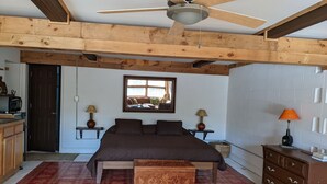 Un dormitorio con una cama grande, vigas de madera en el techo, un ventilador de techo, una ventana, dos mesitas de noche con lámparas, un aparador y un tapete rojo con diseño.