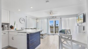 Une cuisine moderne avec des meubles blancs, un mur d’accent bleu, un coin repas avec une table et des chaises blanches, et une vue sur l’extérieur grâce à de grandes fenêtres.