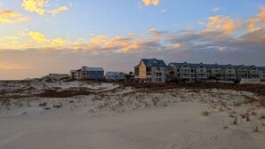 Un hôtel en bord de mer, composé de plusieurs bâtiments, bordé de dunes de sable, et offrant une vue sur un ciel dégagé parsemé de nuages.