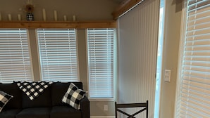 A living room with a dark-colored sofa, a wooden dining table with checkered placemats, and a window with blinds.