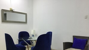 Un petit coin repas avec une table en verre, quatre chaises bleues, une lampe et un canapé en osier agrémenté d’un coussin bleu.
