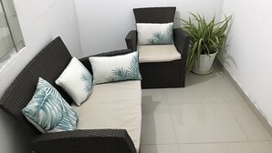 Un coin salon avec un canapé en osier foncé et des fauteuils assortis, chacun agrémenté d’un coussin orné d’un motif de feuilles. On peut apercevoir une plante en pot dans le coin.