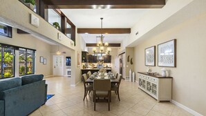 A spacious dining area with a high ceiling, a central chandelier, and a view of the kitchen in the background. - Unique house - heated pool and hot tub option near Clearwater Beach Florida (Clearwater)