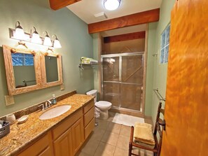 Un baño con tocador doble lavamanos, inodoro, cabina de ducha con mampara de vidrio y una silla de madera.