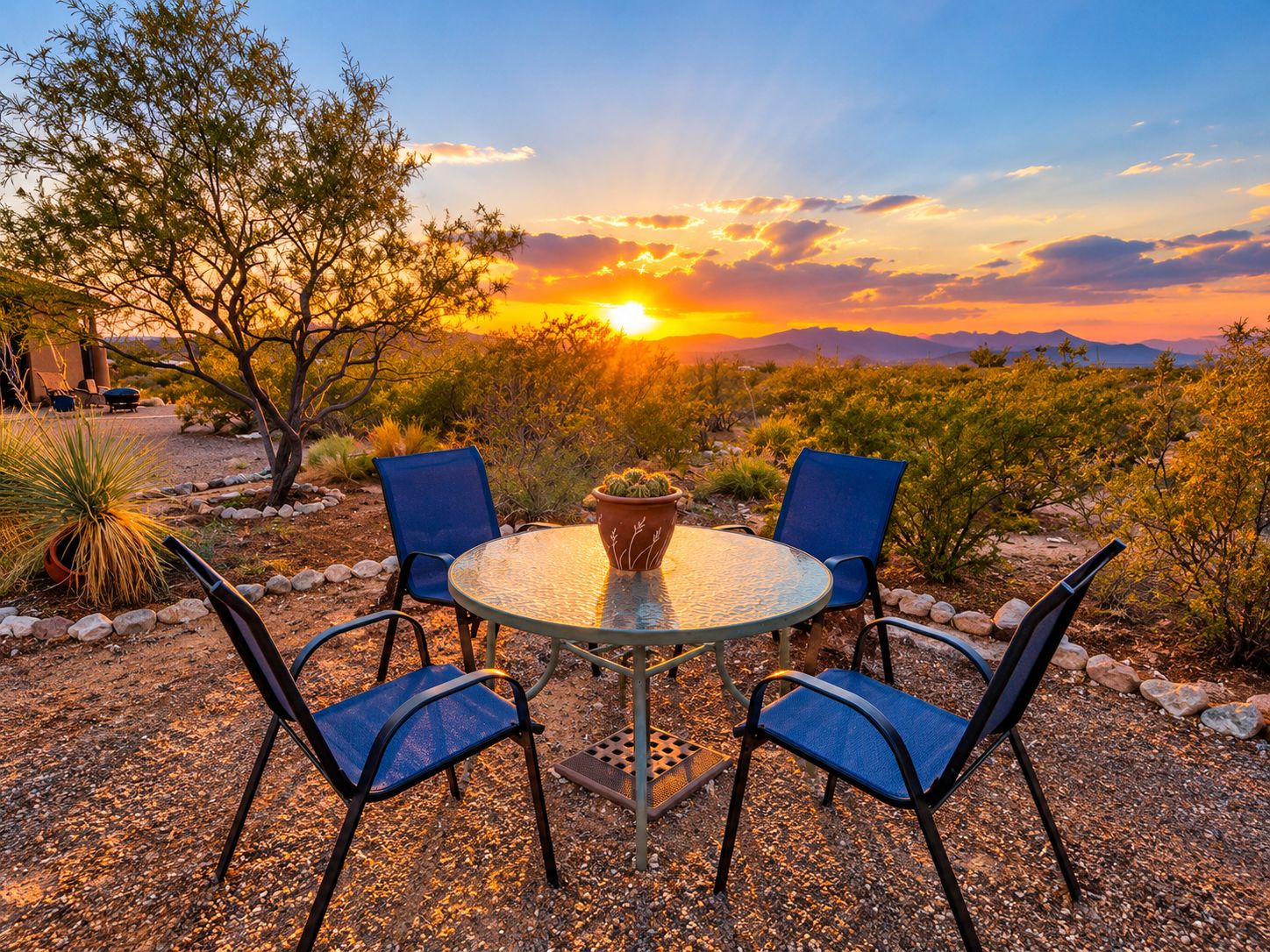 Una mesa de vidrio redonda con cuatro sillas, rodeada de vegetación desértica y una vista al atardecer.