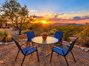 A round glass table with four chairs, surrounded by desert vegetation and a sunset view.