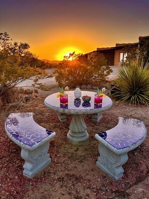 Una mesa redonda con dos sillas, cada una con un asiento de mosaico, ubicada al aire libre con un atardecer de fondo.