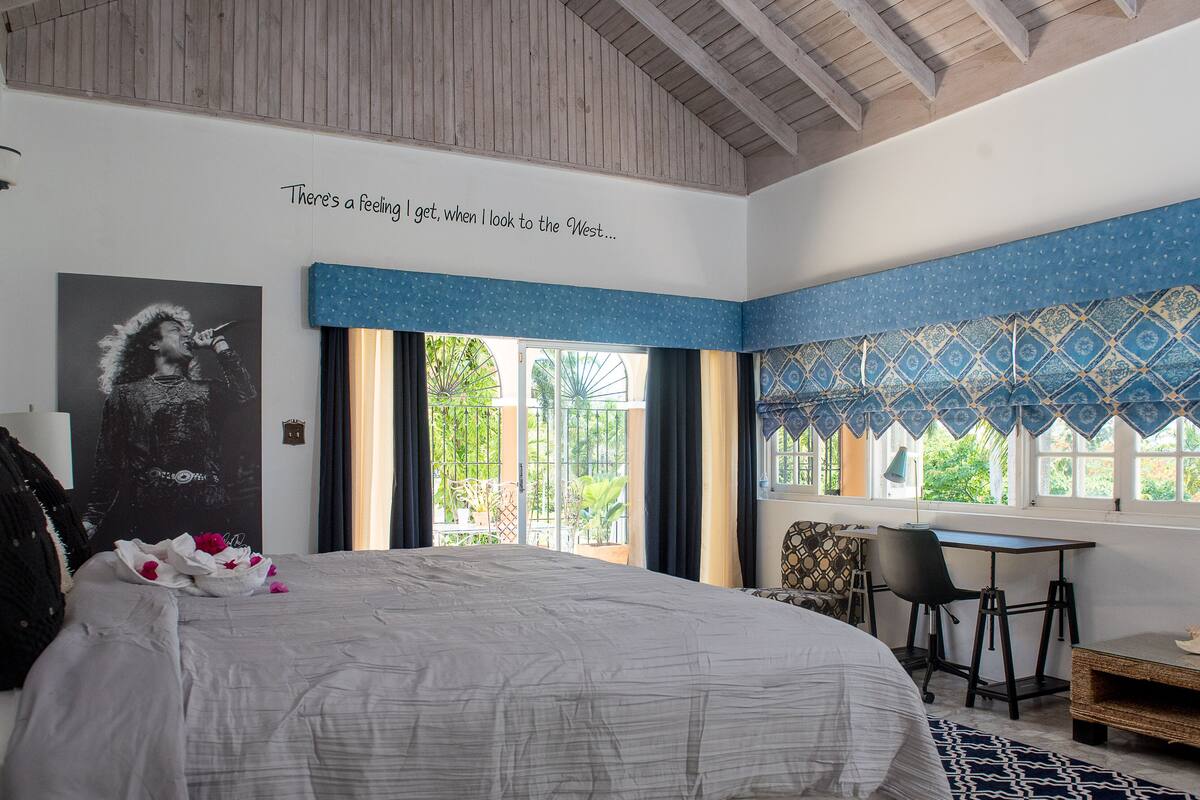 A bedroom with a large bed, a desk, and a chair. There's a window with a view of greenery outside.