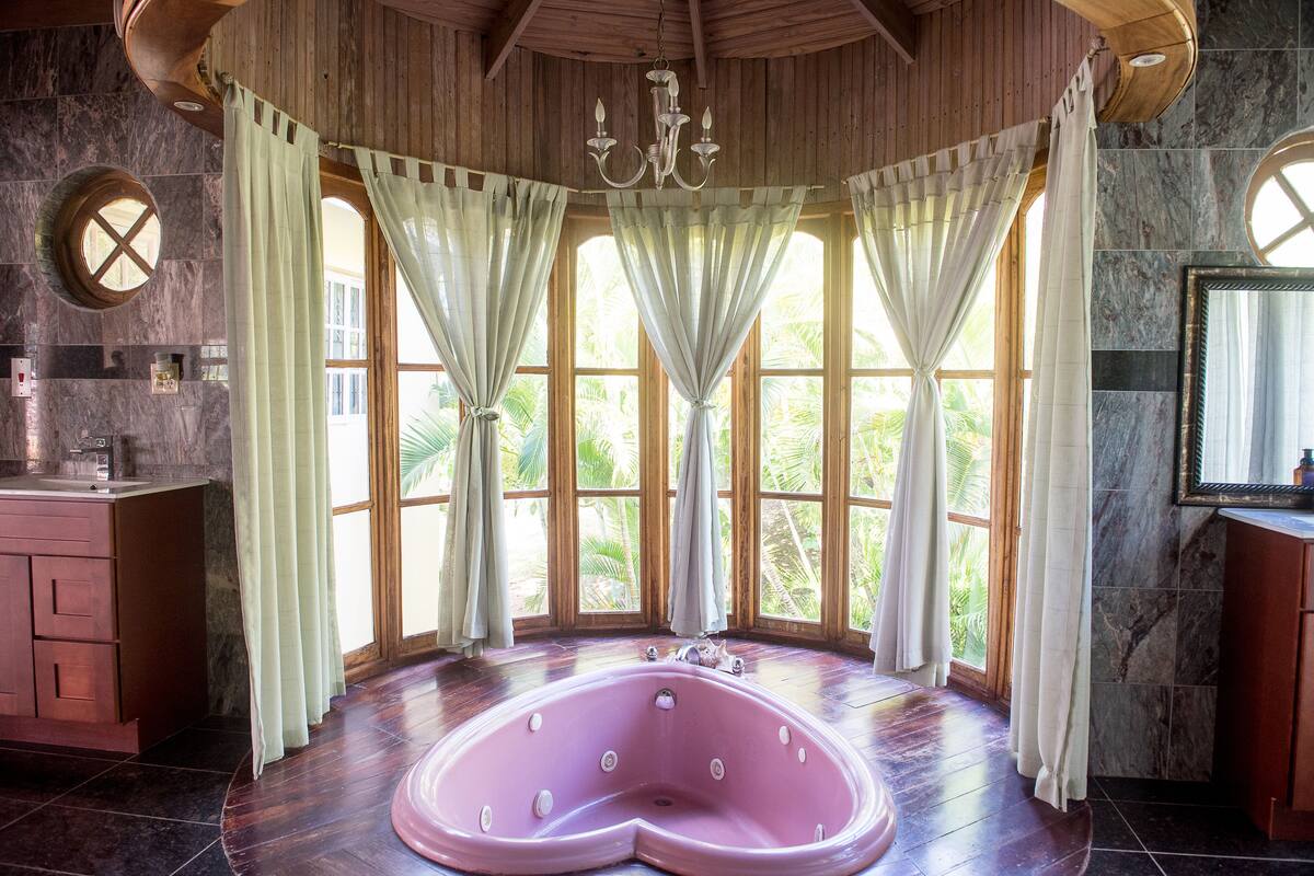A bathroom with a large pink bathtub, wooden ceiling, and large windows with curtains.