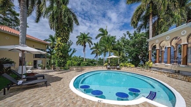 A swimming pool with lounge chairs and umbrellas, surrounded by palm trees and a stone patio. - Music Mansion (Montego Bay)
