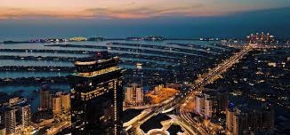 A coastal cityscape at dusk with high-rise buildings, a marina, and a curved road leading to a waterfront area.