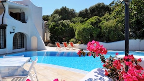 A pool area with a white building, a clear blue pool, and a few lounge chairs.
