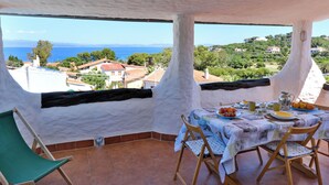 A terrace with a table set for breakfast, a chair, and a view of the sea and hills.