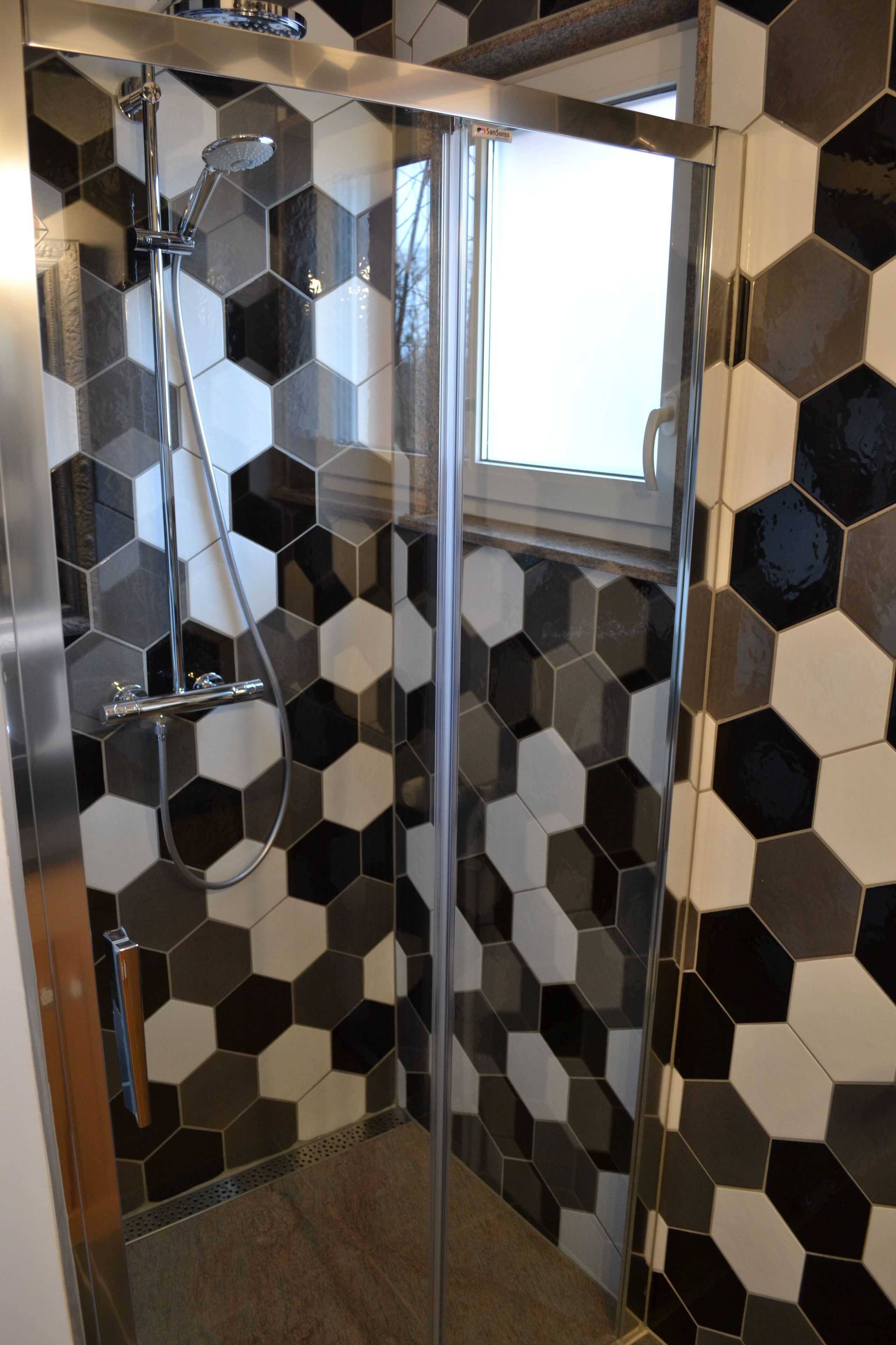 A shower with a glass door, a handheld showerhead, and a wall with a black and white hexagonal tile pattern.