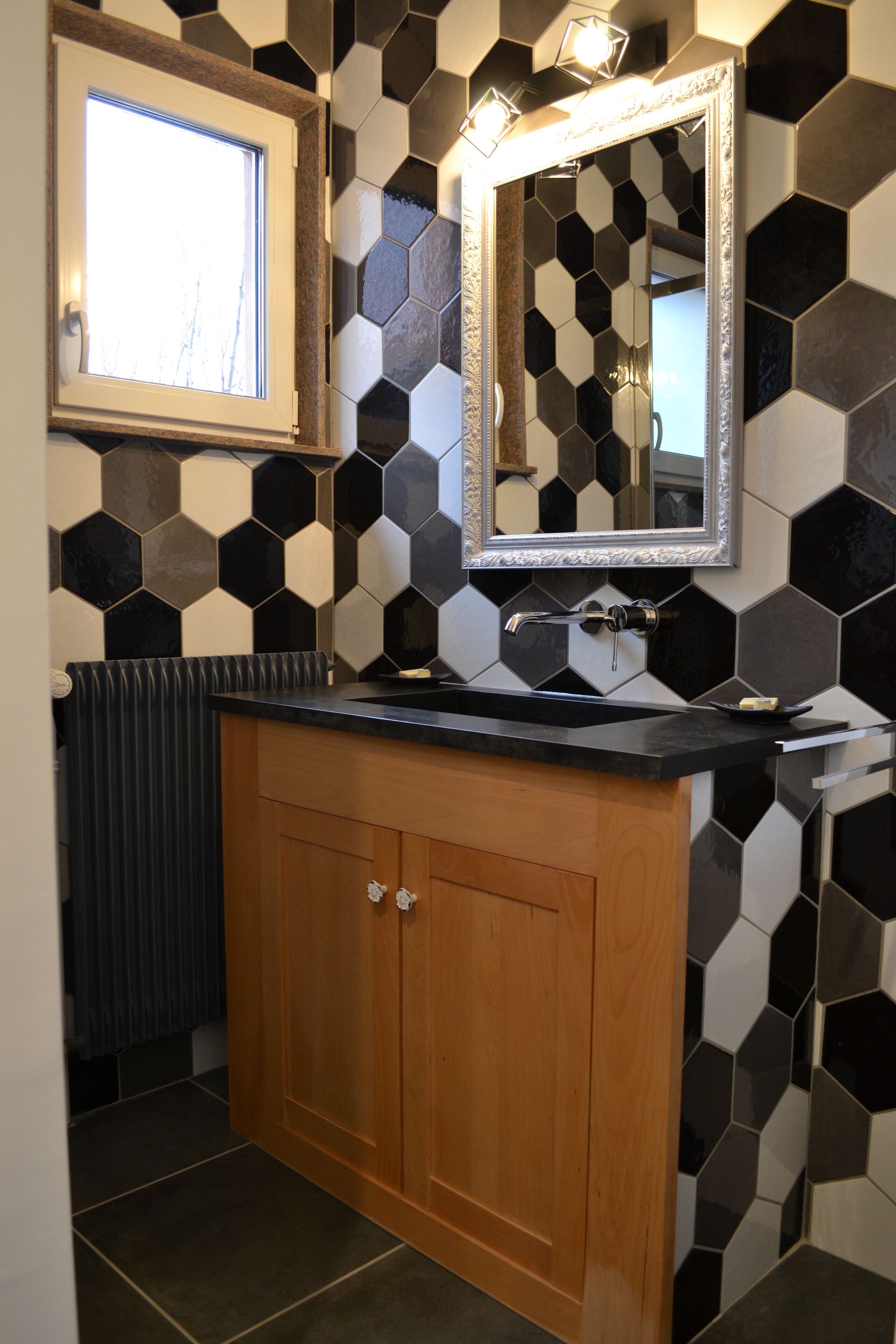 A bathroom with a black and white hexagonal tile backsplash, a wooden vanity with a black countertop, a mirror with an ornate frame, and a window above the sink.